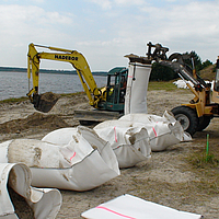 Enchimento com areia disponível localmente dos sacos SoilTain com a ajuda de uma escavadora, enquanto a pá carregadora segura o saco de areia pré-fabricado