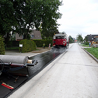 Uma máquina a termo avança, seguida da aplicação do reforço de asfalto SamiGrid® na estrada