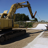 A escavadora utiliza uma travessa de assentamento para estender os tapetes de bentonite Tektoseal® Clay
