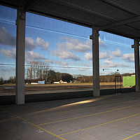 Vista interior das redes corta-vento transparentes num armazém, permitindo a visualização de um campo