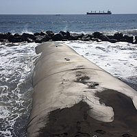 Quebra-mar de geotêxtil construído no mar ao fundo, blocos de água contra os quais as ondas batem